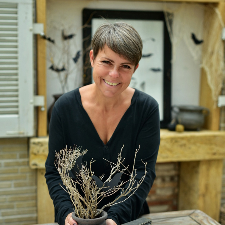 Vrouw in zwart shirt houdt pot met droge takken en vleermuisdecoratie vast in rustieke omgeving