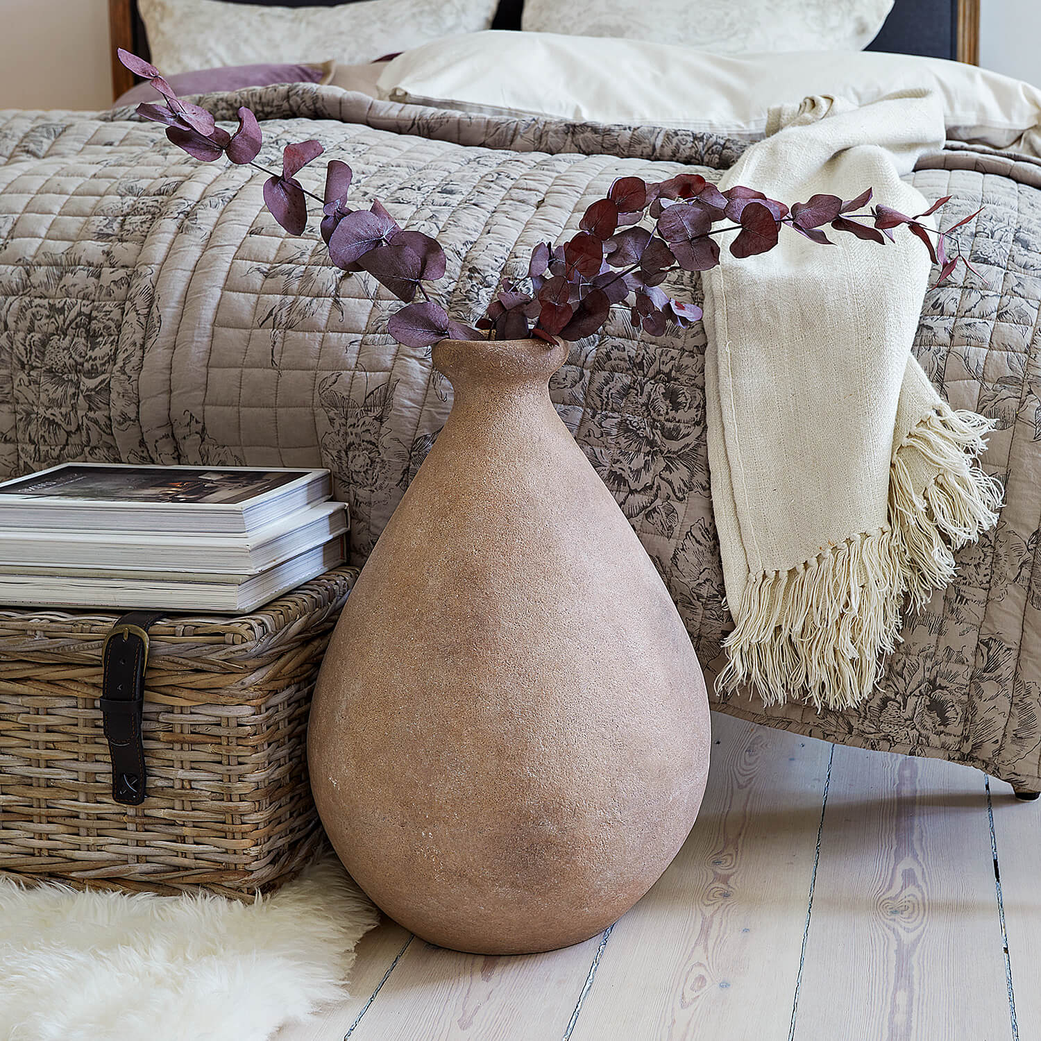 Grote beige keramische vaas met gedroogde takken voor bed en rieten mand