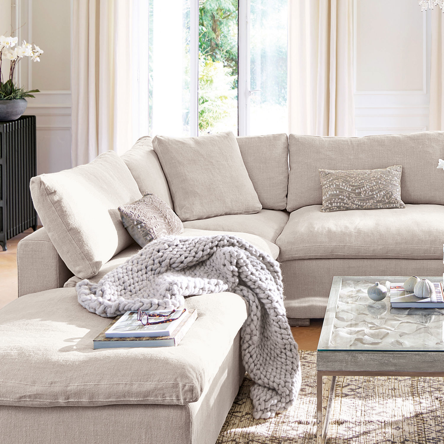 Beige stoffen hoekbank met kussens en grijze gebreide deken in lichte woonkamer