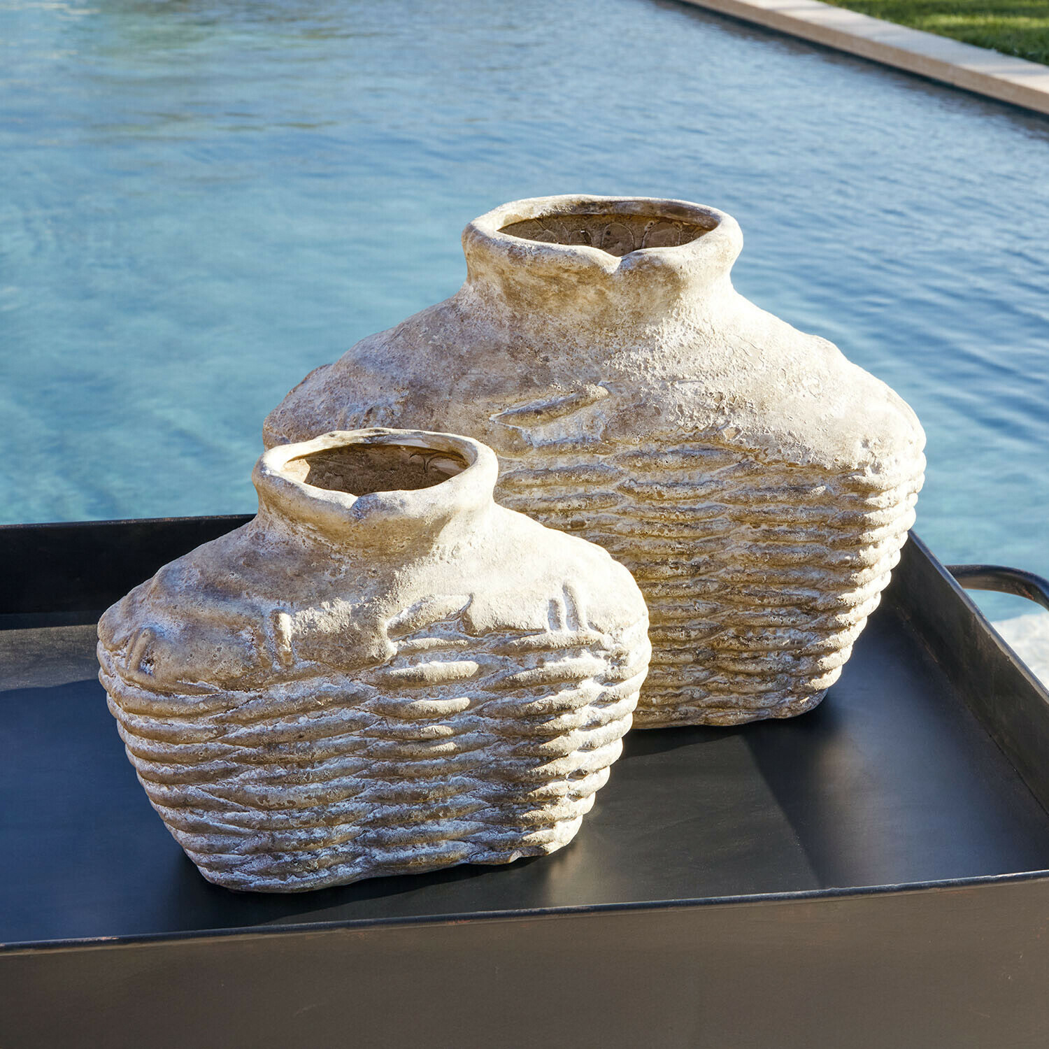 Twee decoratieve beige stenen vazen op een zwart dienblad bij een zwembad