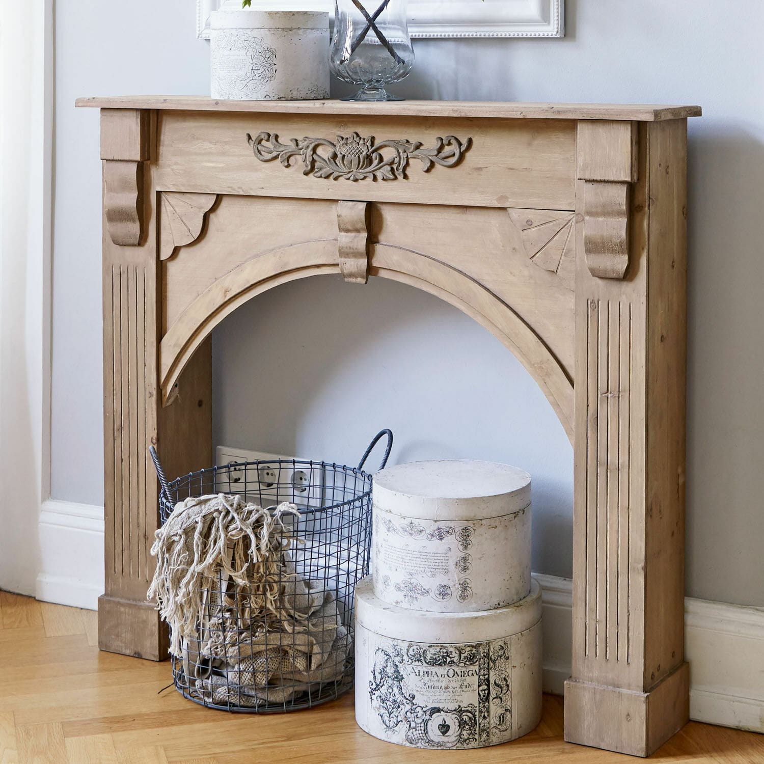 Houten schoorsteenmantel met gesneden patroon in natuurlijke kleur, metalen mand en decoratieve witte dozen op de vloer