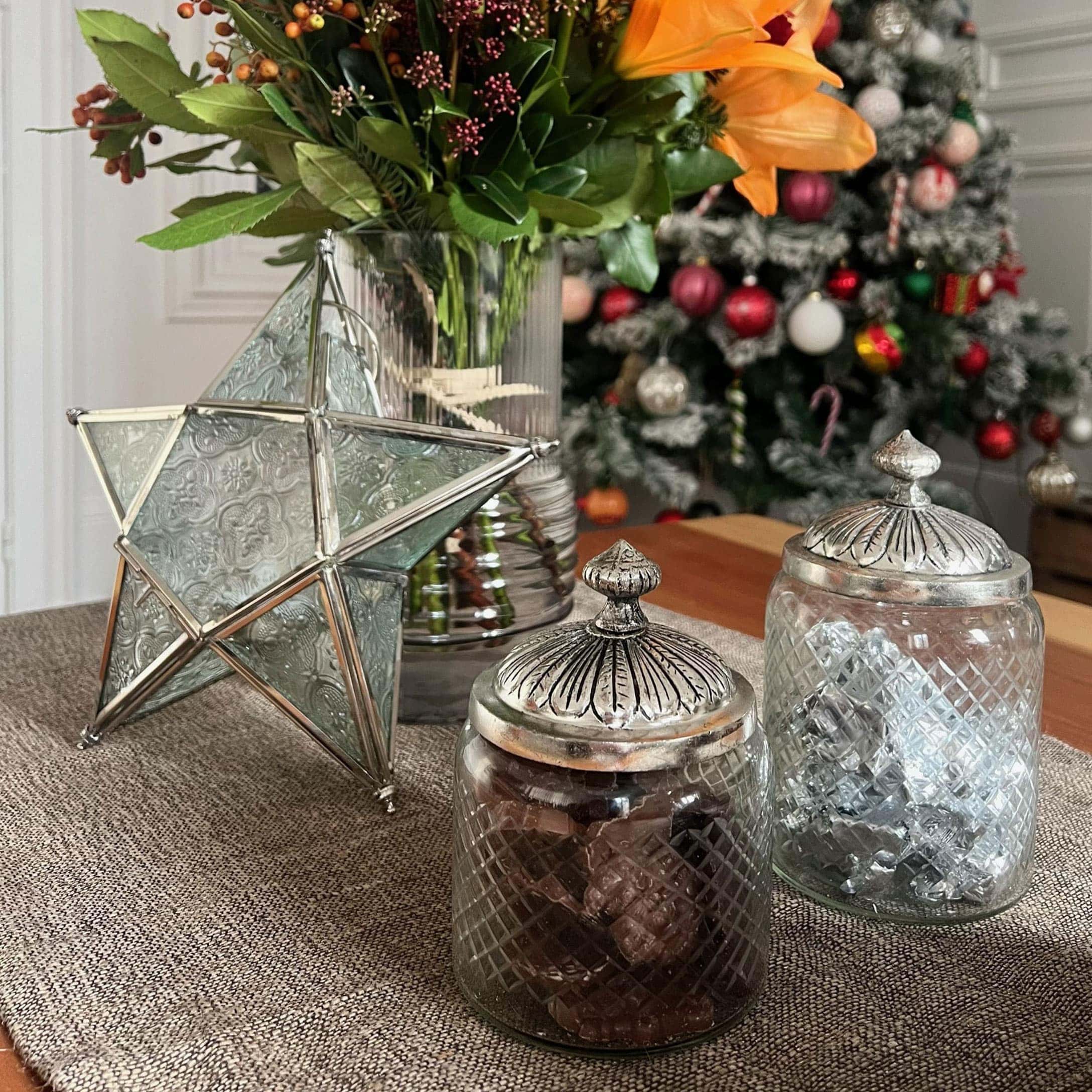 Glazen potten met metalen deksels en decoratieve ster op tafel met bloemen en kerstboom op de achtergrond