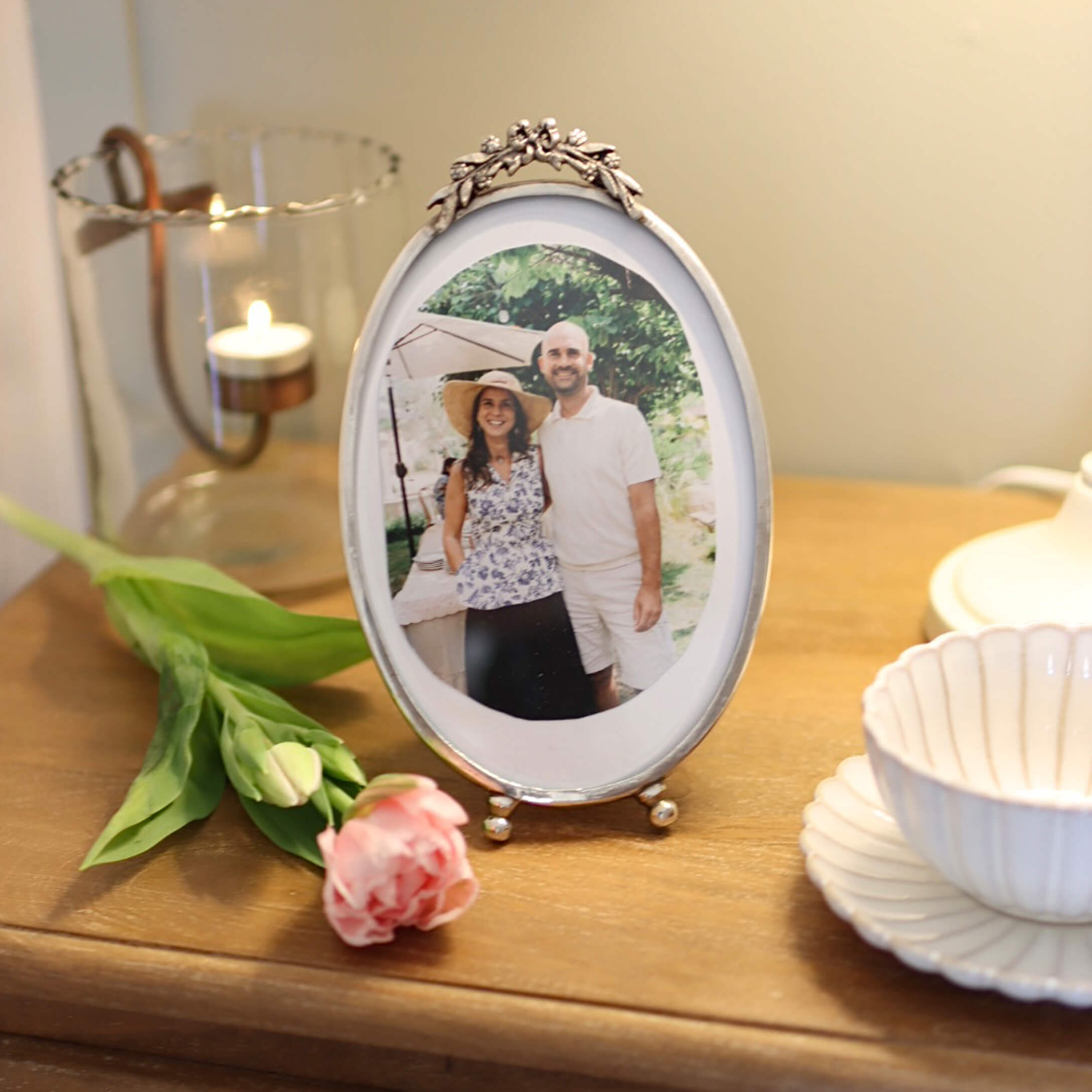 Ovale zilveren fotolijst met bloemmotief op houten tafel met roos en windlicht