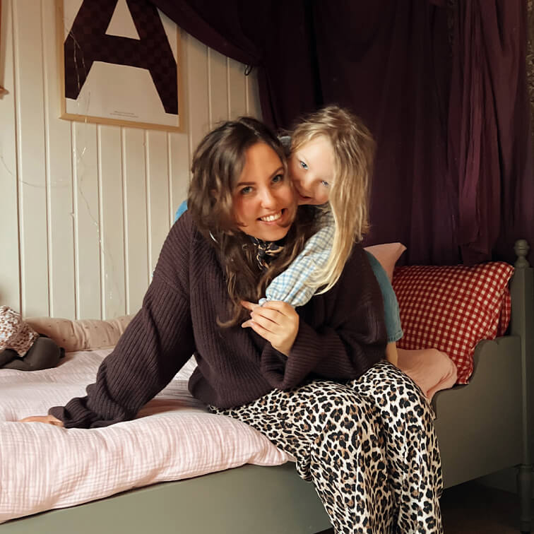 Vrouw in bruine trui en luipaardbroek zittend op bed met kind in licht geruit overhemd