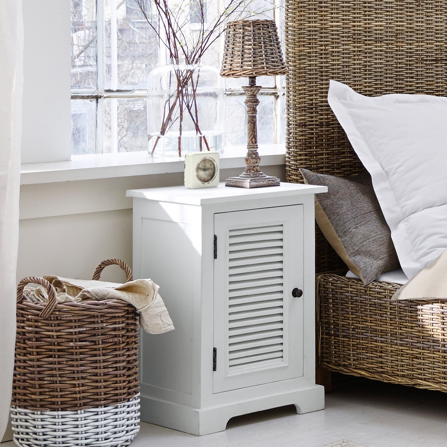 Witte houten nachtkastje met luikdeur naast rotan bed en wasmand in slaapkamer