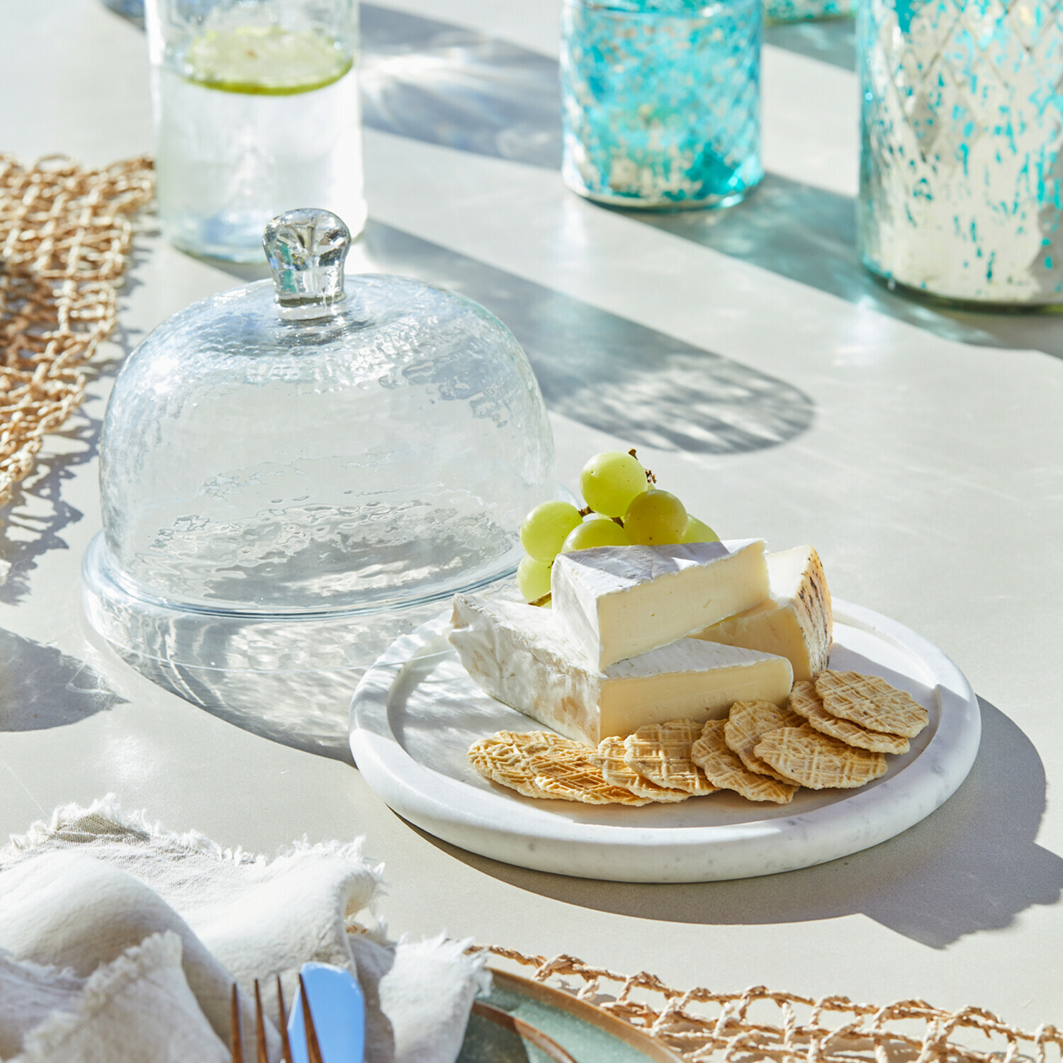 Transparante glazen stolp naast bord met kaas, crackers en groene druiven op lichte tafel