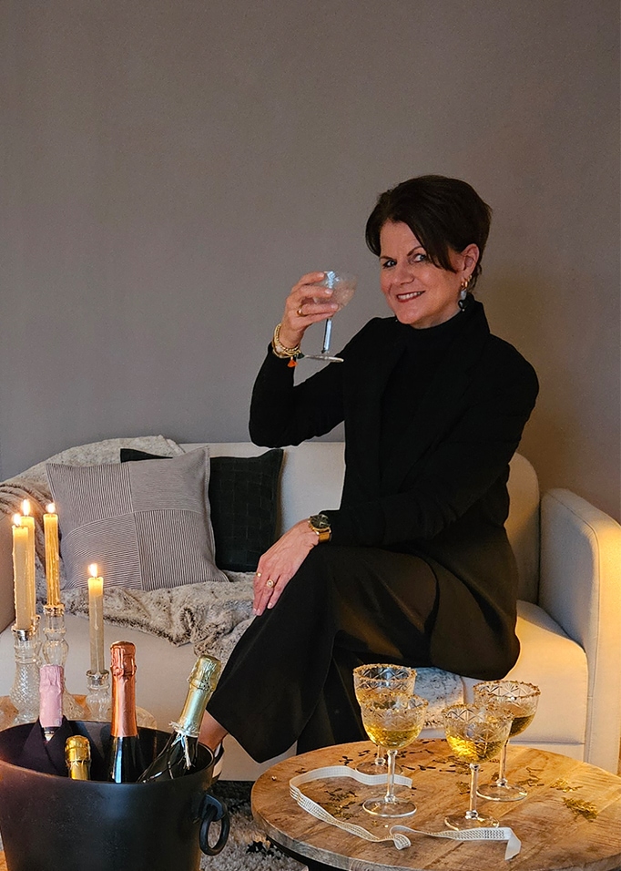 Vrouw in zwarte kleding zittend op lichte bank met glas, kaarsen en champagneflessen ernaast