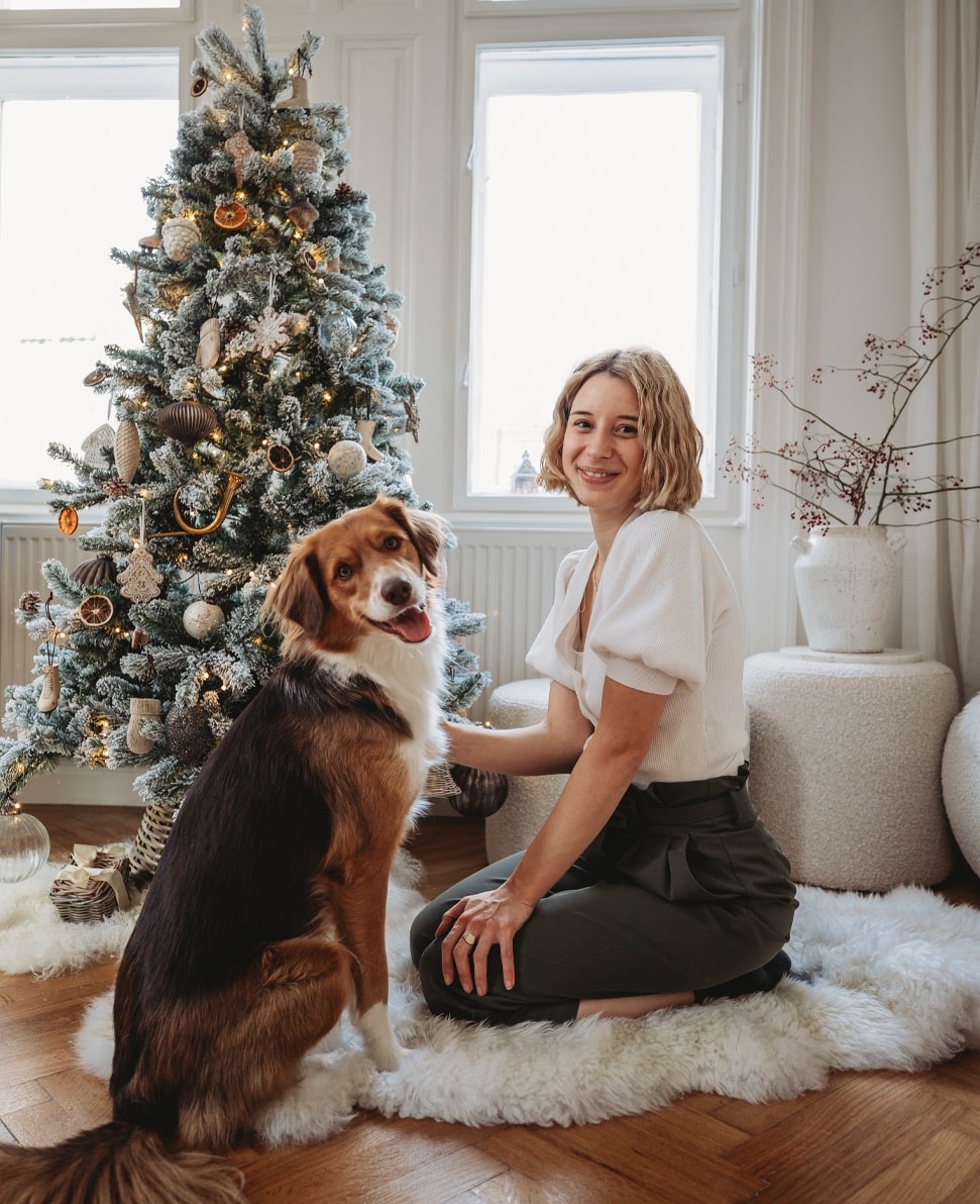Vrouw in wit shirt en groene broek met hond naast versierde kerstboom in lichte kamer
