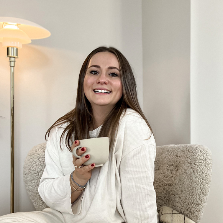 Lachende vrouw in witte trui met beige mok, zittend in zachte stoel in lichte kamer