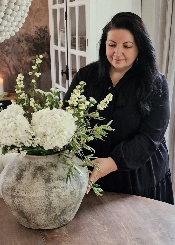 Vrouw die een wit boeket schikt in een grote grijze keramische vaas op een houten tafel