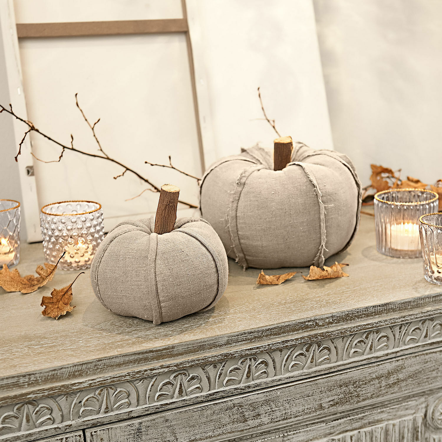 Decoratieve pompoenen van beige linnen met houten steeltjes op een sierlijke kast met windlichten