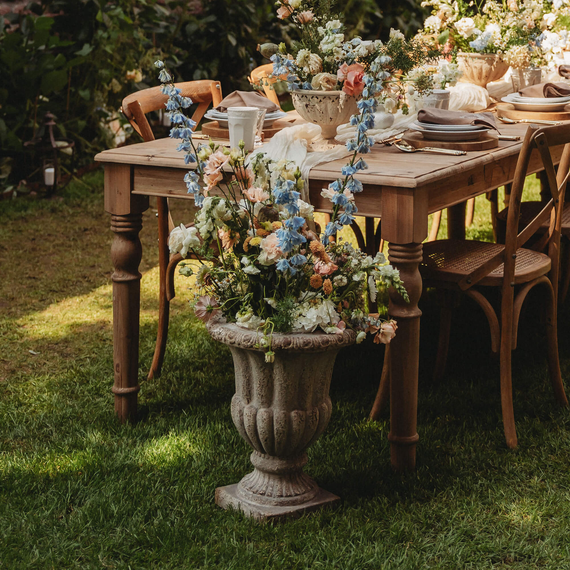 Grote stenen vaas met pastelkleurige bloemen voor houten tafel en stoelen in groene tuin