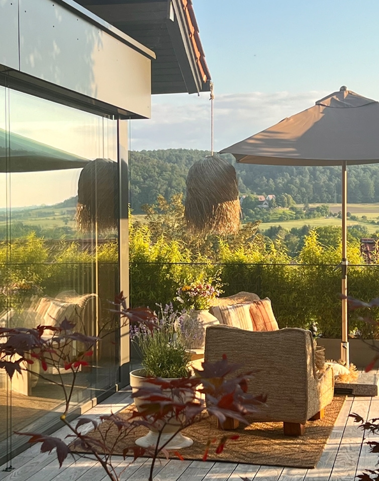 Gezellig terras met beige bank, parasol en planten tegen een groene landschap en heldere lucht.