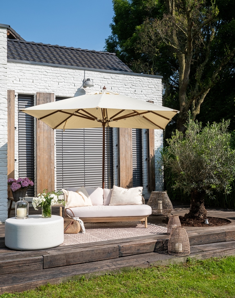 Terras met witte bakstenen muur, beige bank met kussens en crèmekleurige parasol op houten terras