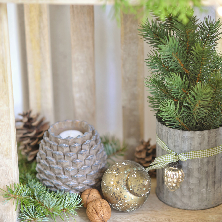 Kaarsenhouder in dennenappelvorm met groene takken in metalen pot op houten plank