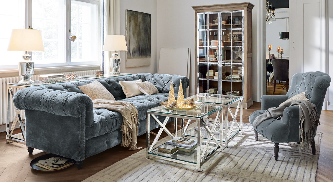 Grijze fluwelen Chesterfield bank en stoel met knoopdetails in stijlvolle woonkamer met metalen glazen tafels.