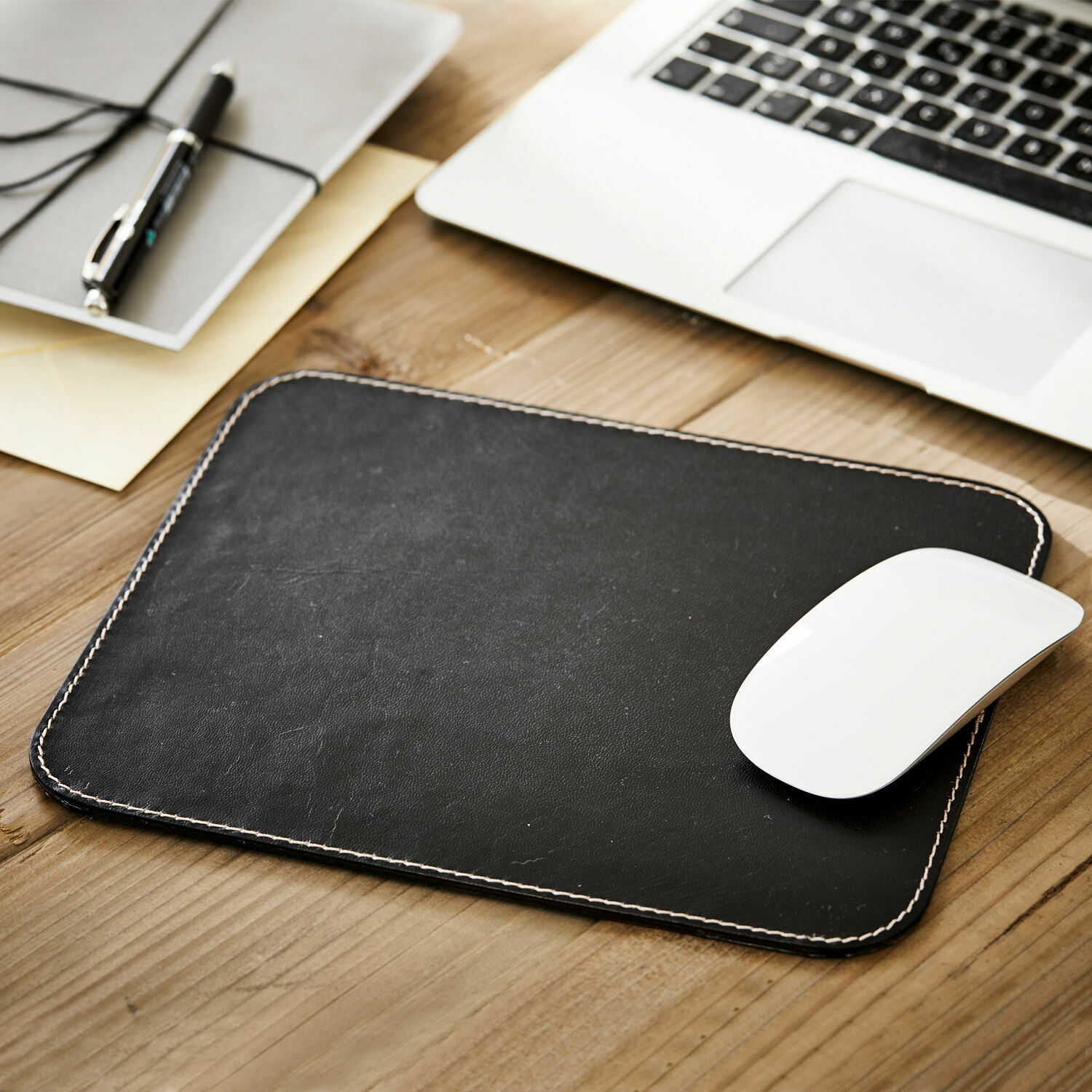 Zwarte leren muismat met witte stiksels op houten bureau naast laptop