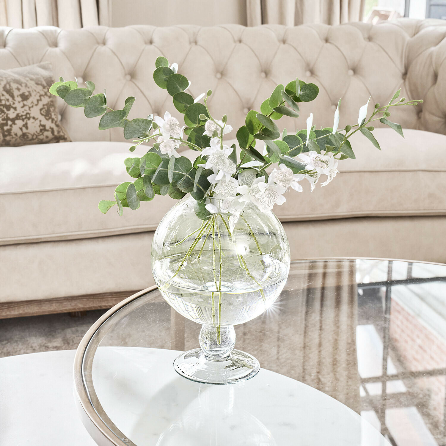 Vaas Marsetta Transparante glazen vaas met groene bladeren en witte bloemen op glazen tafel in lichte woonkamer