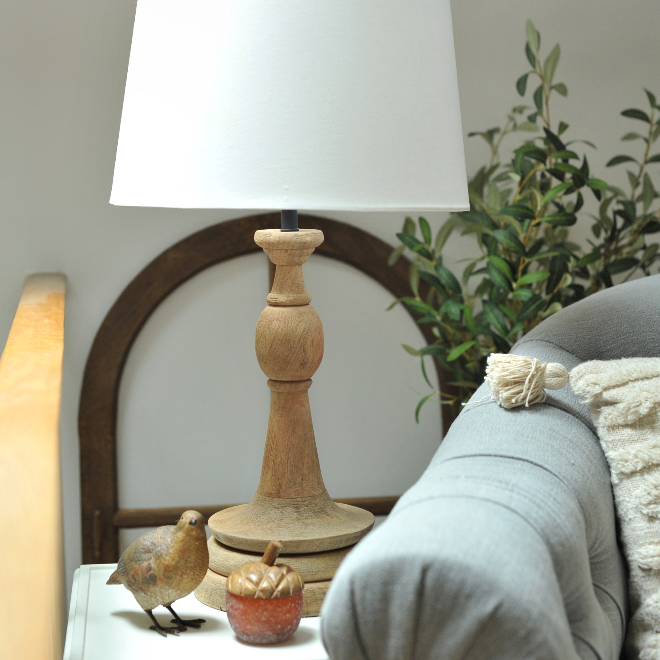 Tafellamp met houten voet en witte kap op tafel naast grijze bank en decoraties