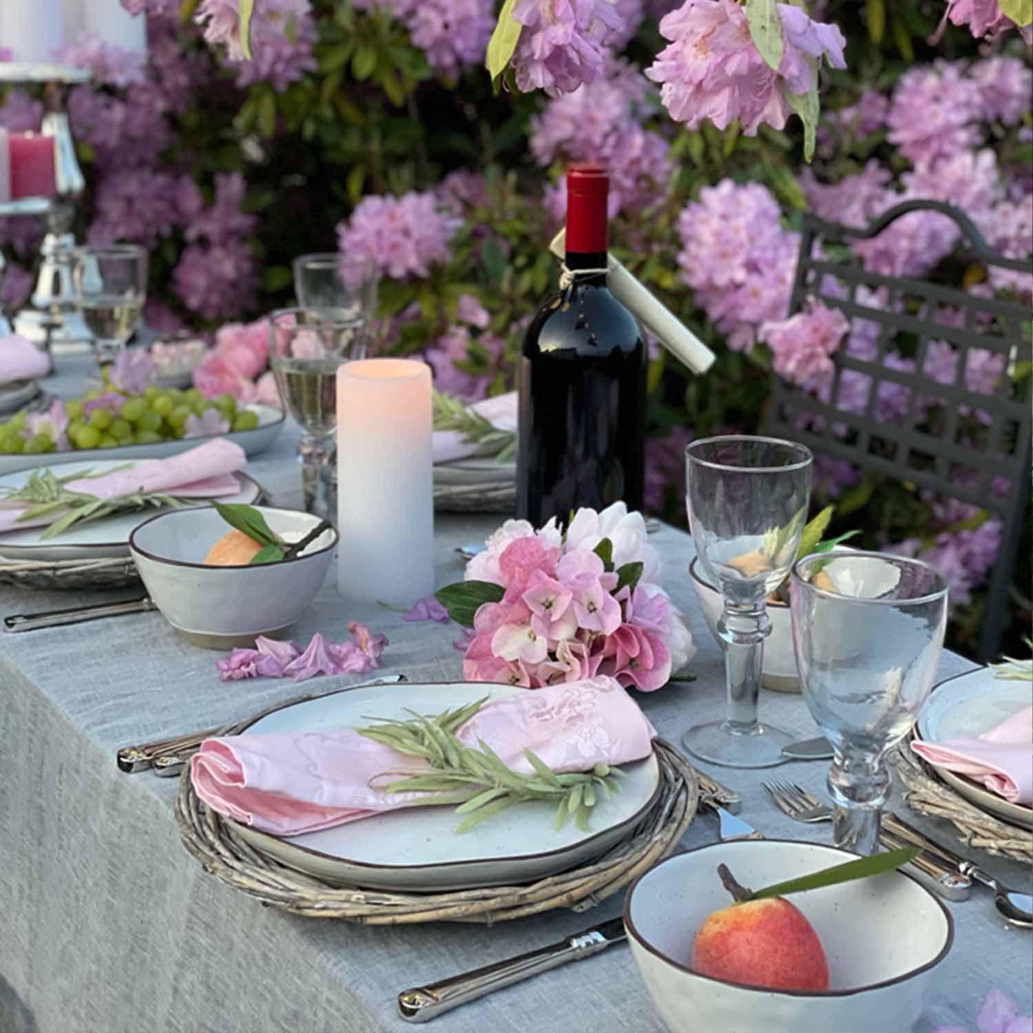 Stijlig gedekte tafel met wit servies, roze servetten en fles rode wijn in bloeiende tuin