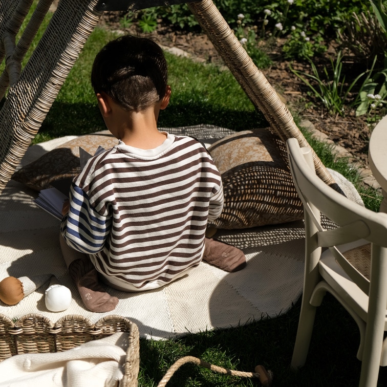 Kind in gestreepte trui zit op tapijt onder rieten tent in tuin, ontspannen speelmoment