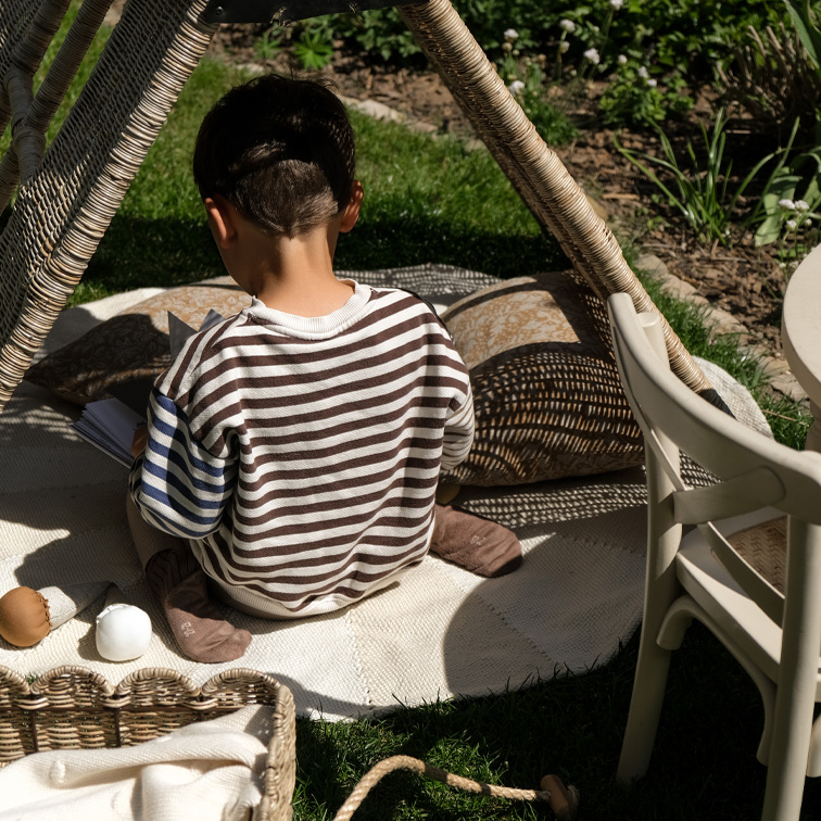 Kind in gestreepte trui zit op tapijt onder rieten tent in tuin, ontspannen speelmoment