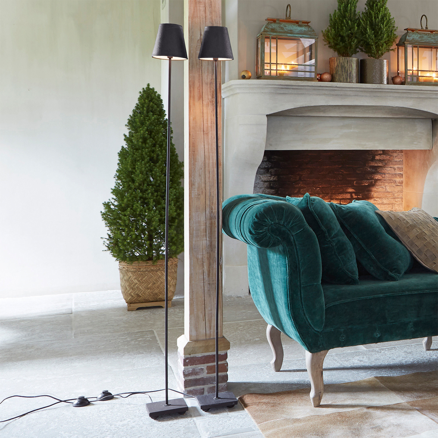 Donkergroene fluwelen bank met kussens naast open haard en zwarte vloerlampen in woonkamer