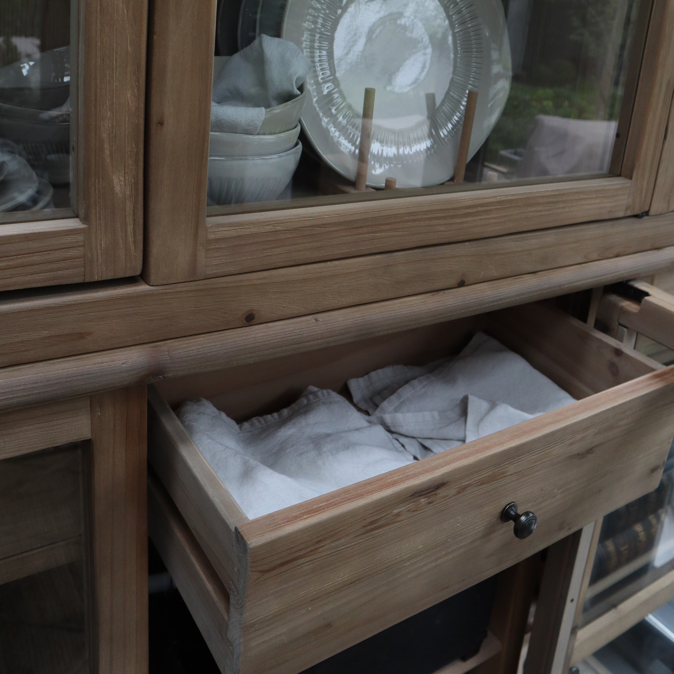 Houten kast met open lade en witte linnen doeken, keramisch servies zichtbaar achter glas