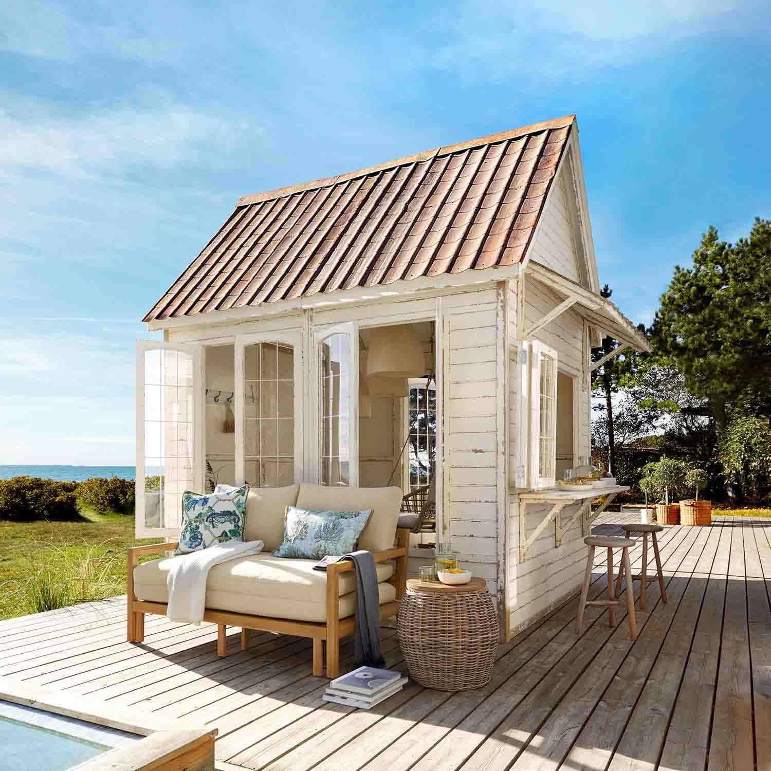 Witte houten tuinhuis met rood dak op houten terras met beige bank en rieten tafel