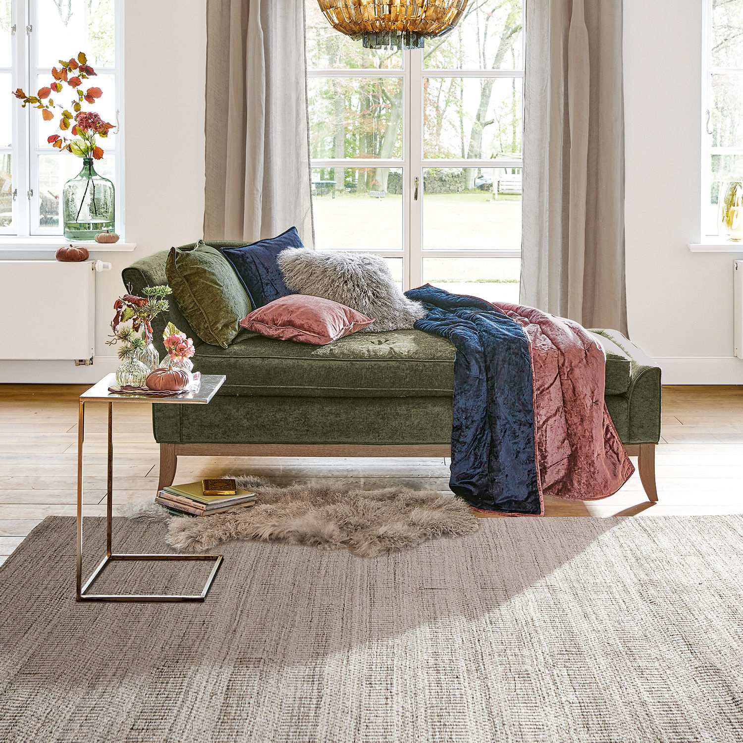 Groene fluwelen chaise longue met kussens en roze-blauwe deken in lichte woonkamer met groot raam