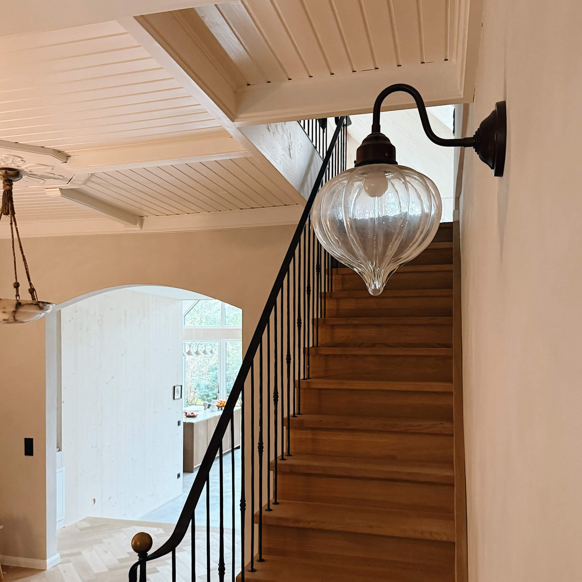 Glazen wandlamp met zwart metalen arm boven houten trap in lichte ruimte