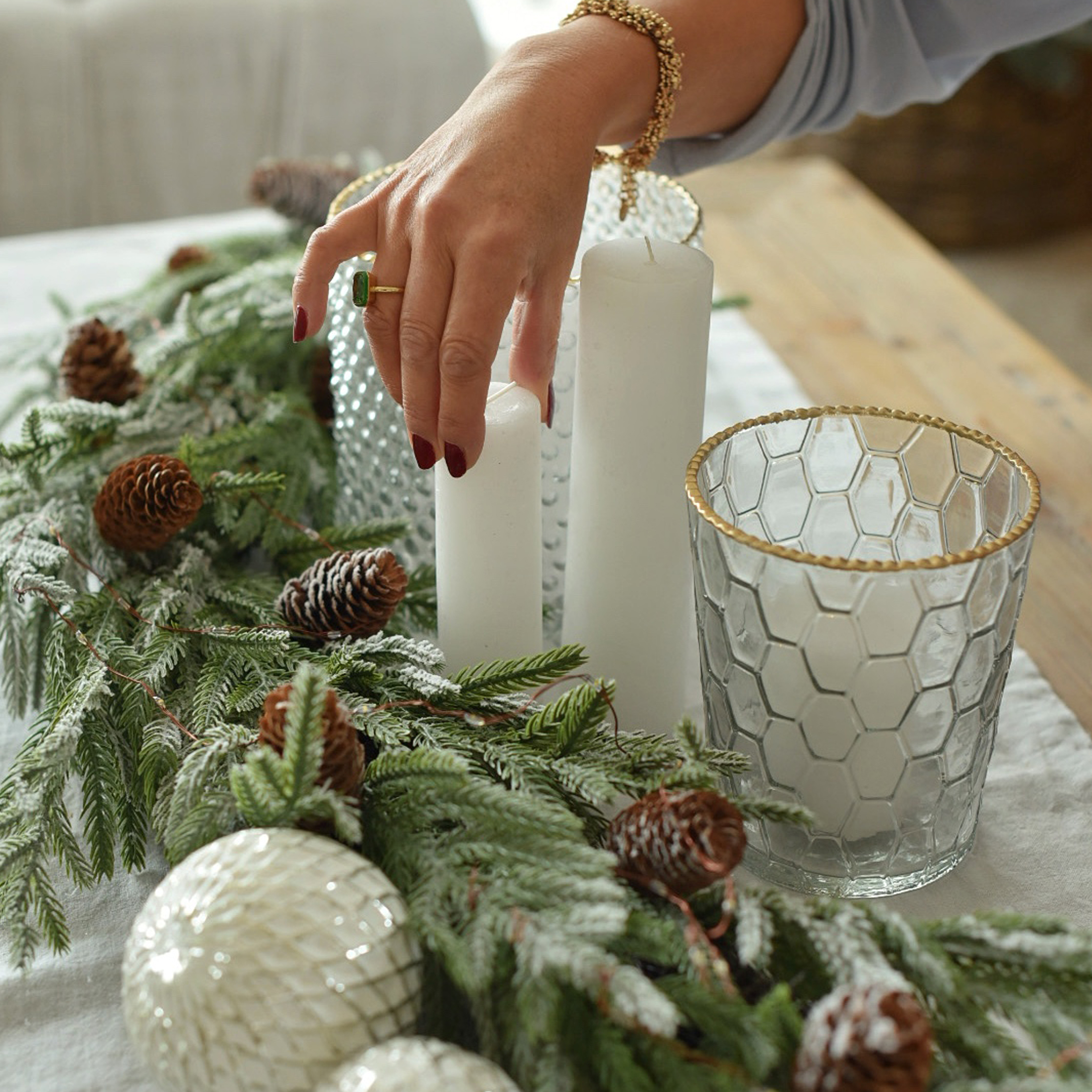 Kerstdecoratie met witte kaarsen, glazen windlicht met gouden rand en groene takken met dennenappels