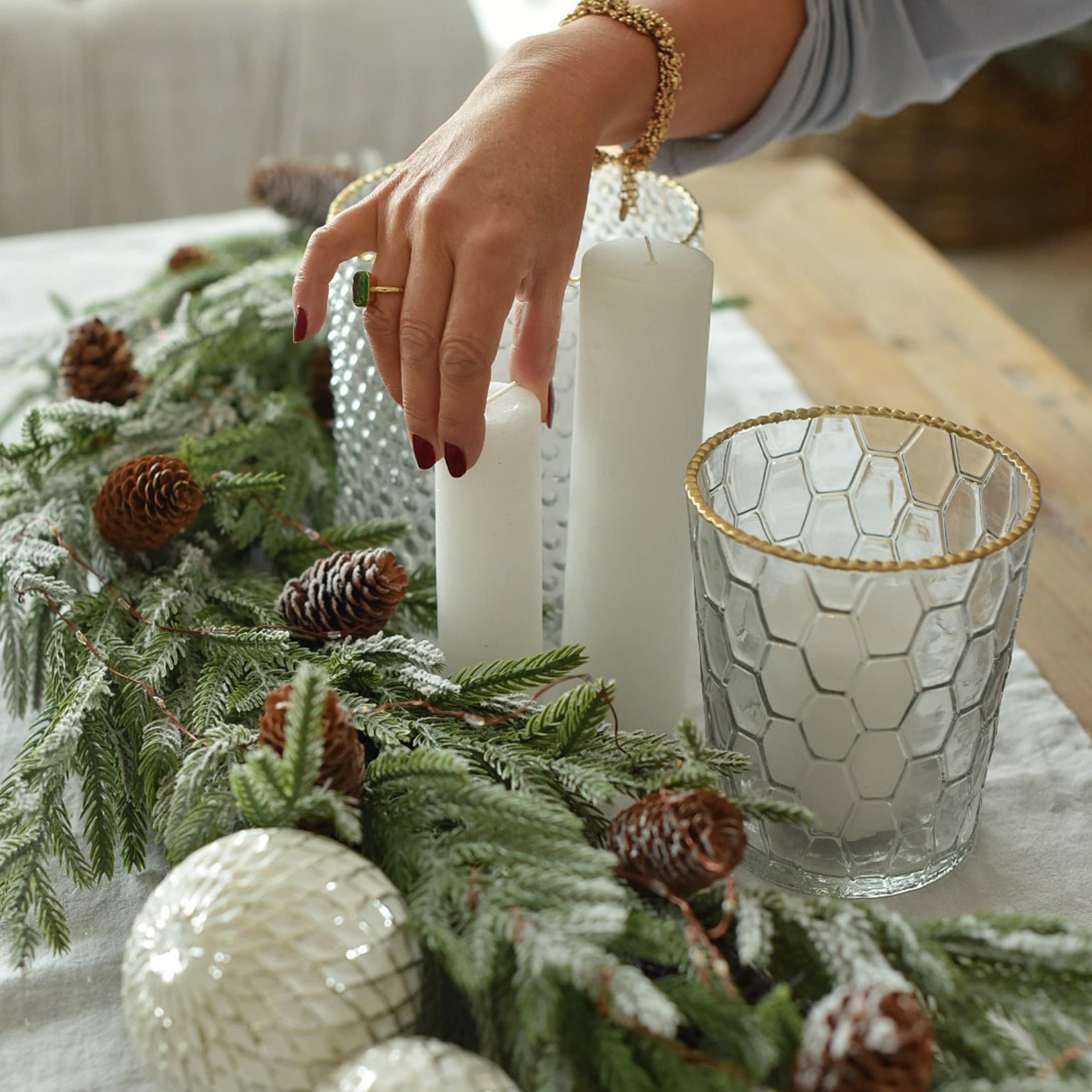 Kerstdecoratie met witte kaarsen, glazen windlicht met gouden rand en groene takken met dennenappels