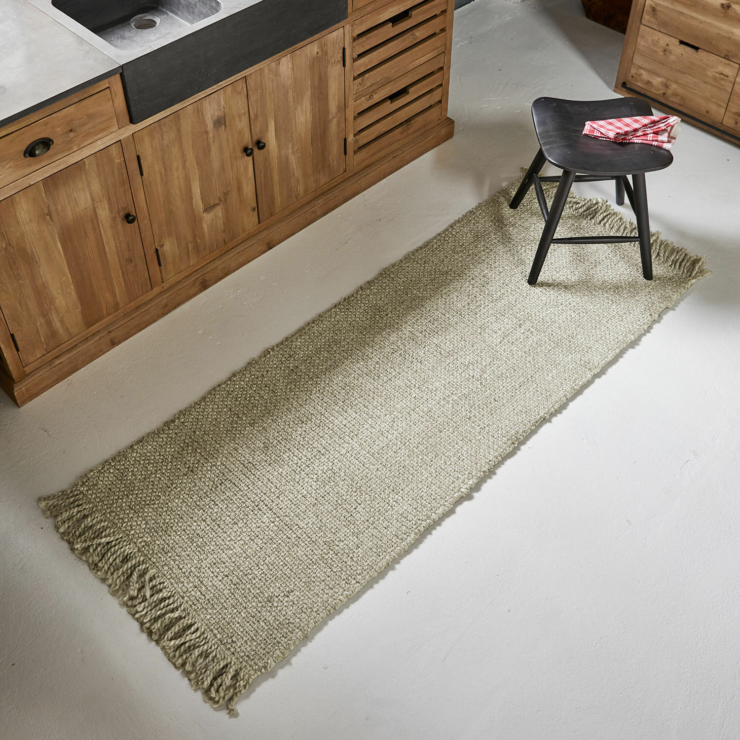 Lange beige vloerkleed met franjes op keukenvloer naast houten kast en zwarte kruk