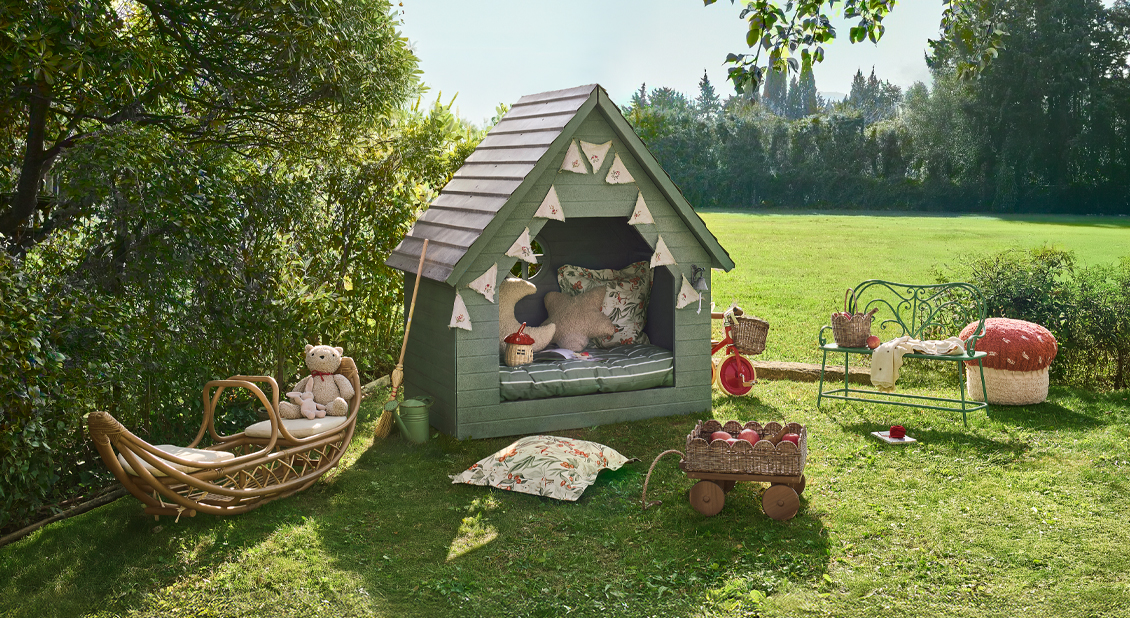 Groen houten speelhuisje met kussens en speelgoed op gras in zonnige tuin