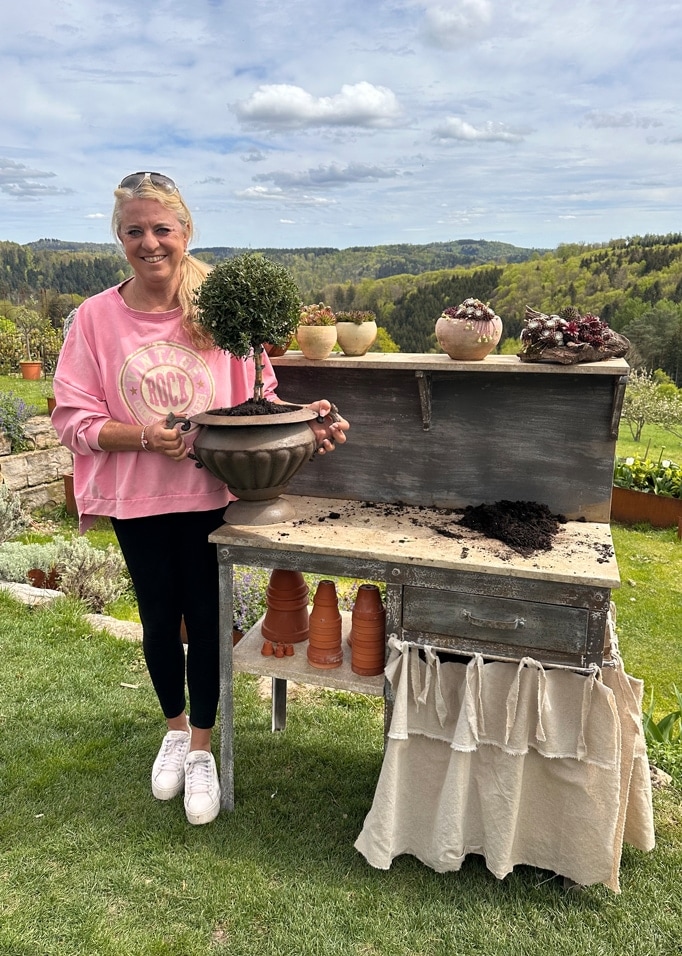 Vrouw in roze trui houdt pot met groene struik vast bij houten tuintafel met groene landschap op achtergrond