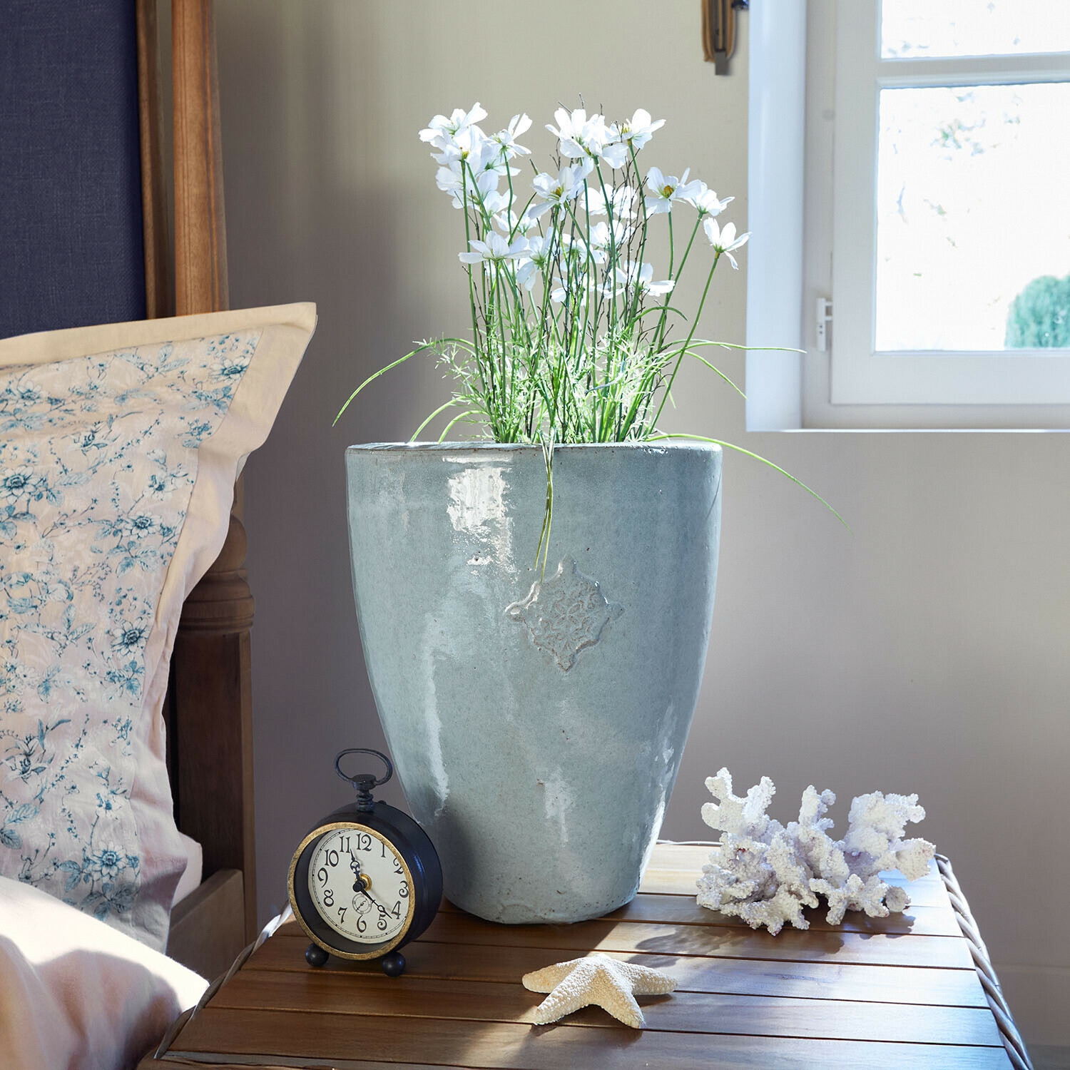 Sierpot Serenzia Grijze keramische bloempot met witte bloemen op houten tafel naast wekker en zee decoraties
