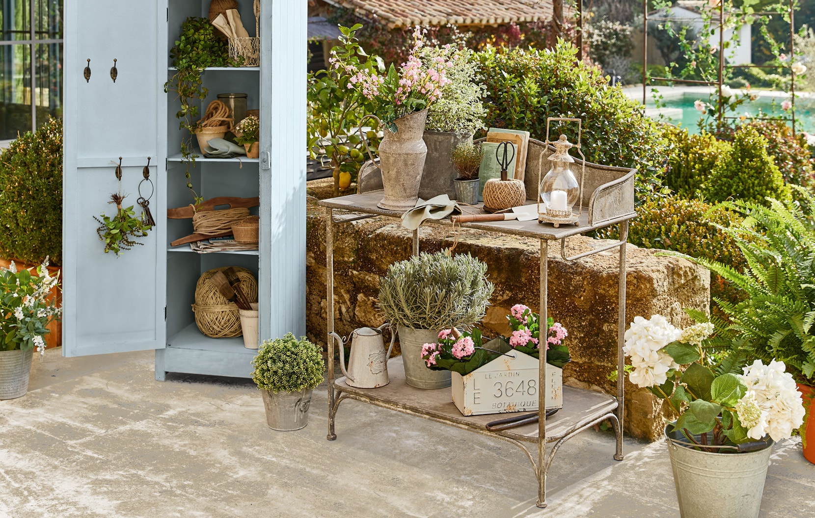 Metalen tuinwagen met shabby chic decoraties en bloemen op terras bij blauwe kast.