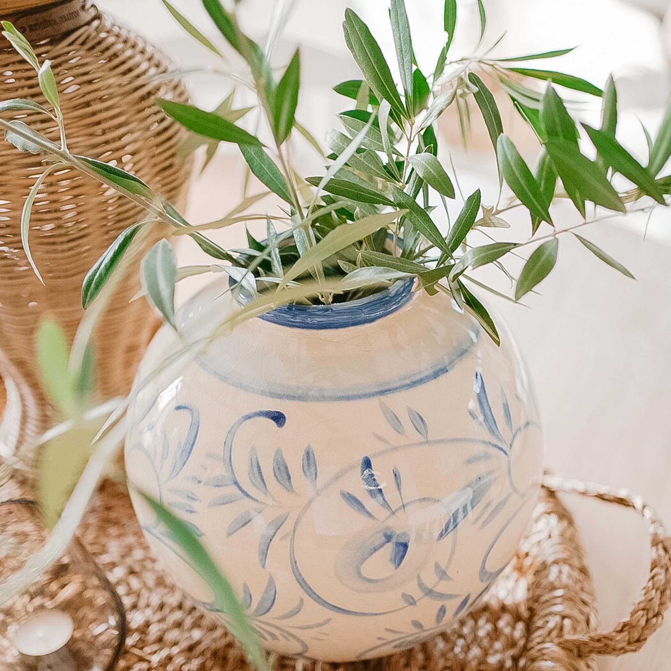 Witte keramische vaas met blauw patroon en groene bladeren op een gevlochten dienblad, lentebloemstuk