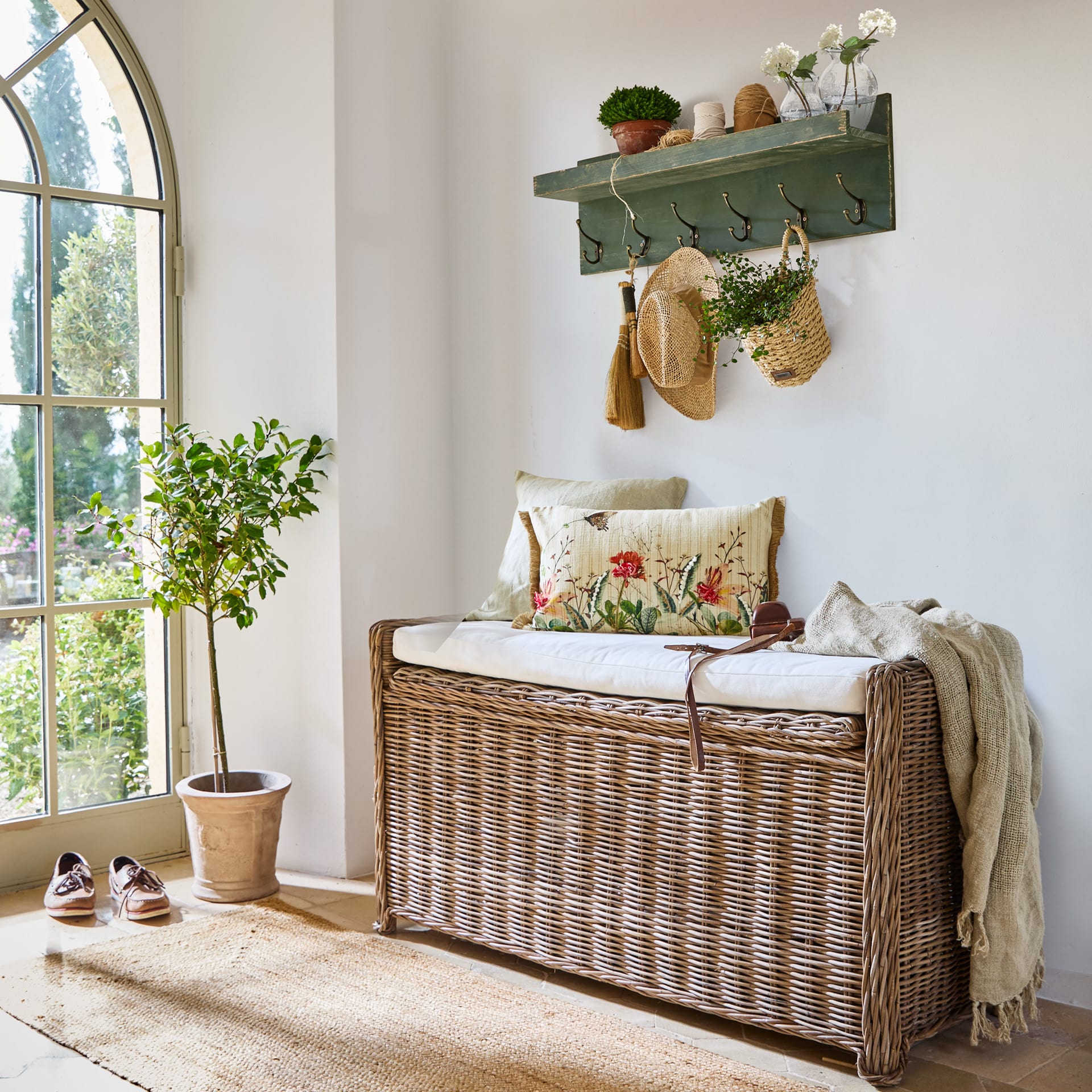 Rotan bank met wit kussen en decoratieve kussens in lichte kamer met groene kapstok