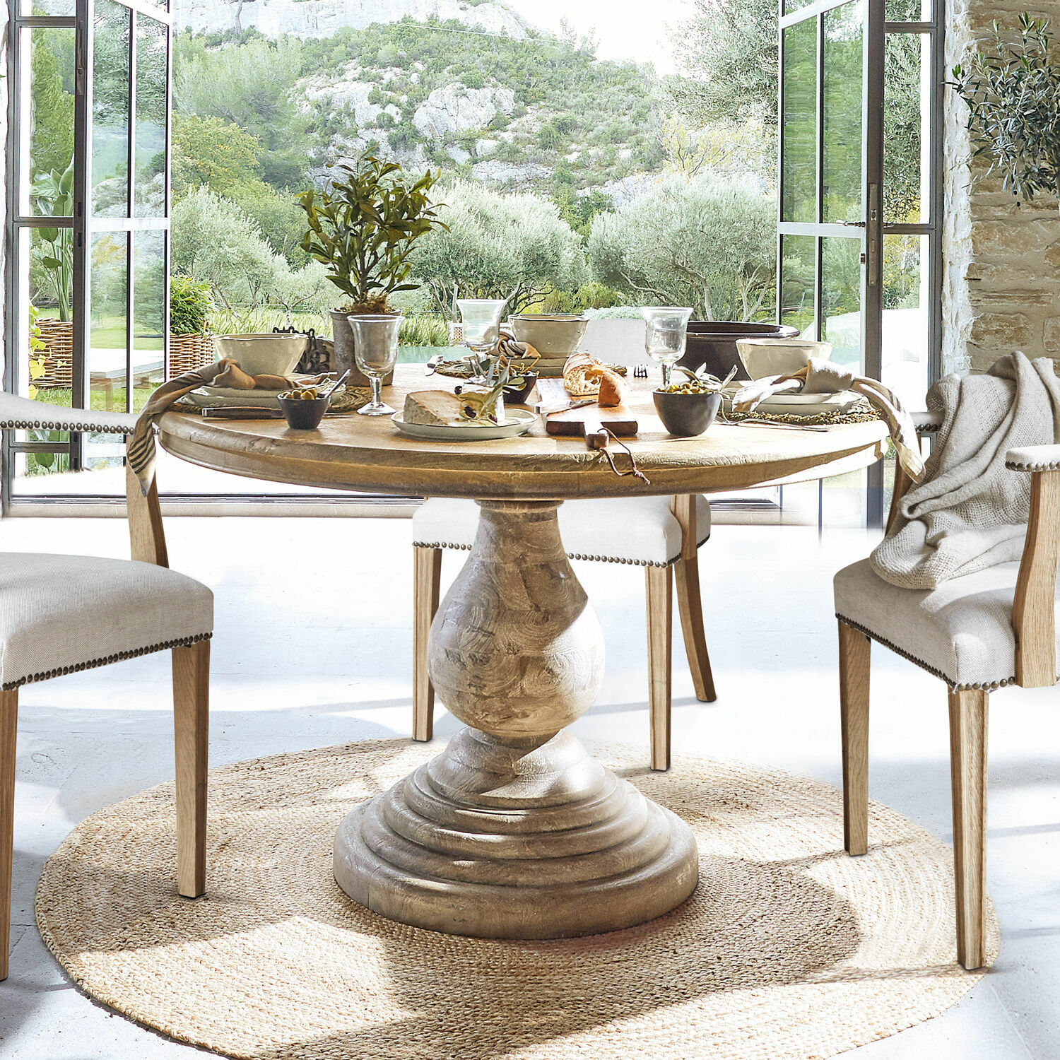 Ronde houten tafel met sierlijke voet op natuurlijke vloerkleed in lichte eetkamer met uitzicht op tuin