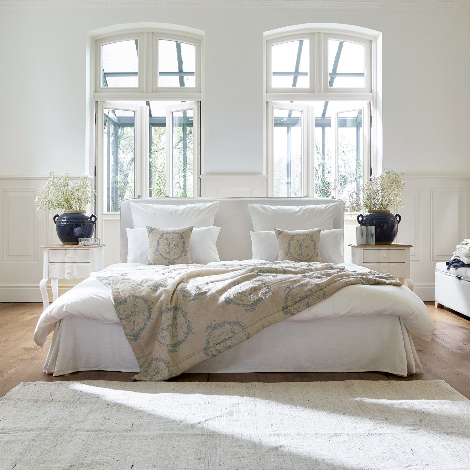 Bed Aubrianne Gezellig bed met lichte bekleding en beige deken met patroon in lichte slaapkamer met grote ramen