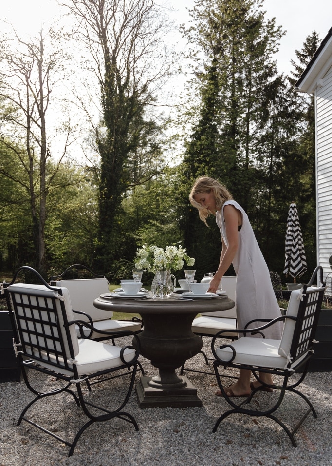 Tuin met ronde stenen tafel en zwarte metalen stoelen met witte kussens, vrouw in witte jurk.