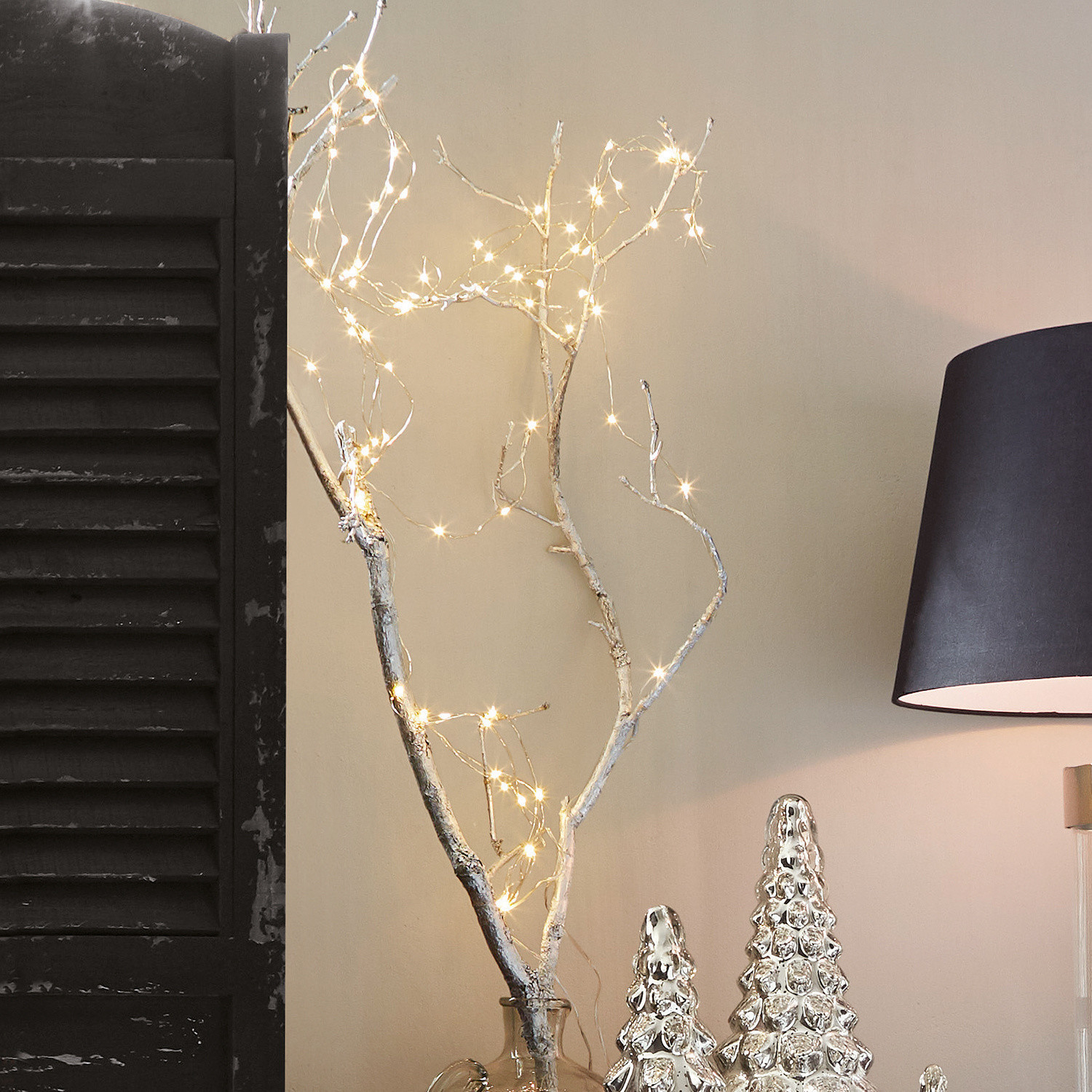 Lichtsnoer Loperhet Decoratieve tak met LED-lichtjes in een glazen vaas naast zilveren kerstdecoraties en een zwarte lamp