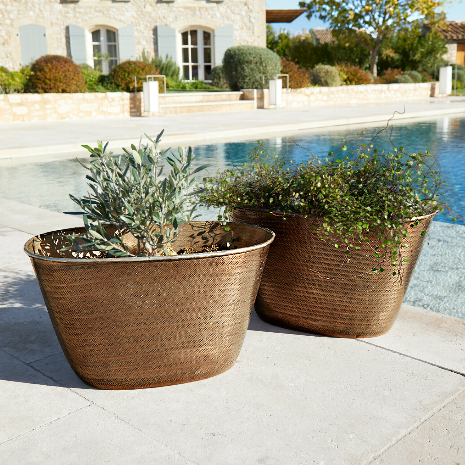 Twee koperen plantenbakken met planten naast een zwembad op een stenen terras, tuindecoratie