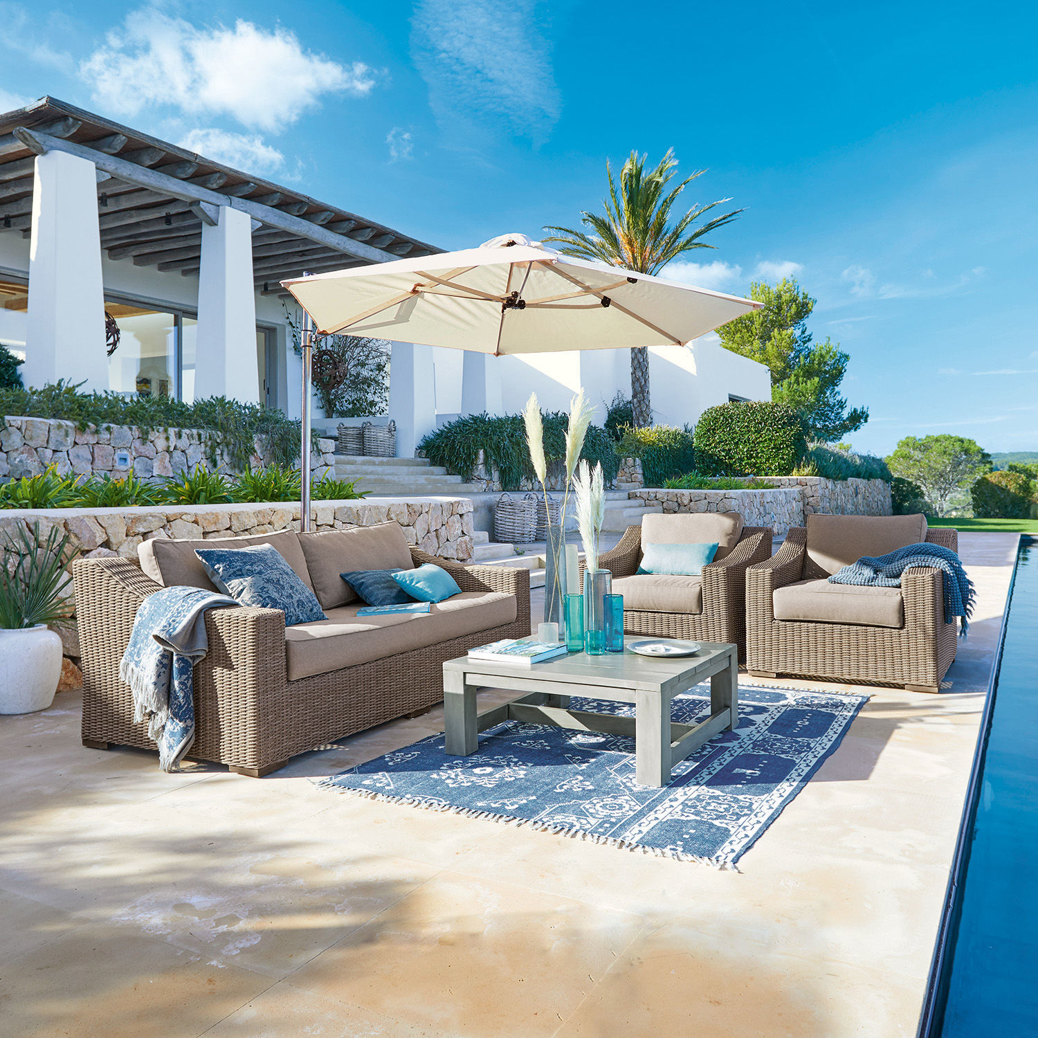Zitgroep Chestingdale Beige rotan tuinmeubelset met parasol en blauw tapijt op zonnig terras