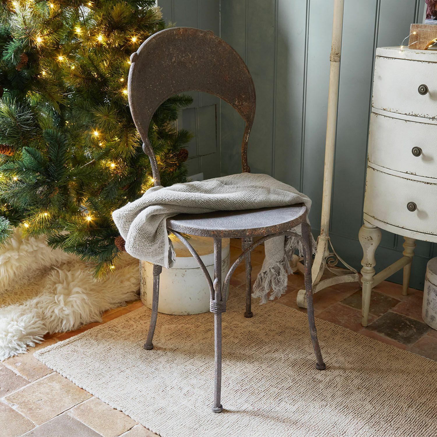 Rustieke metalen stoel met roestkleur en grijze deken bij verlichte kerstboom en beige vloerkleed