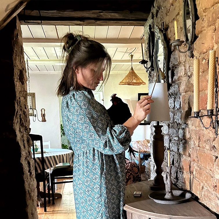 Vrouw in blauw patroonjurk steekt lamp aan in rustiek interieur met bakstenen muur en kaarsen