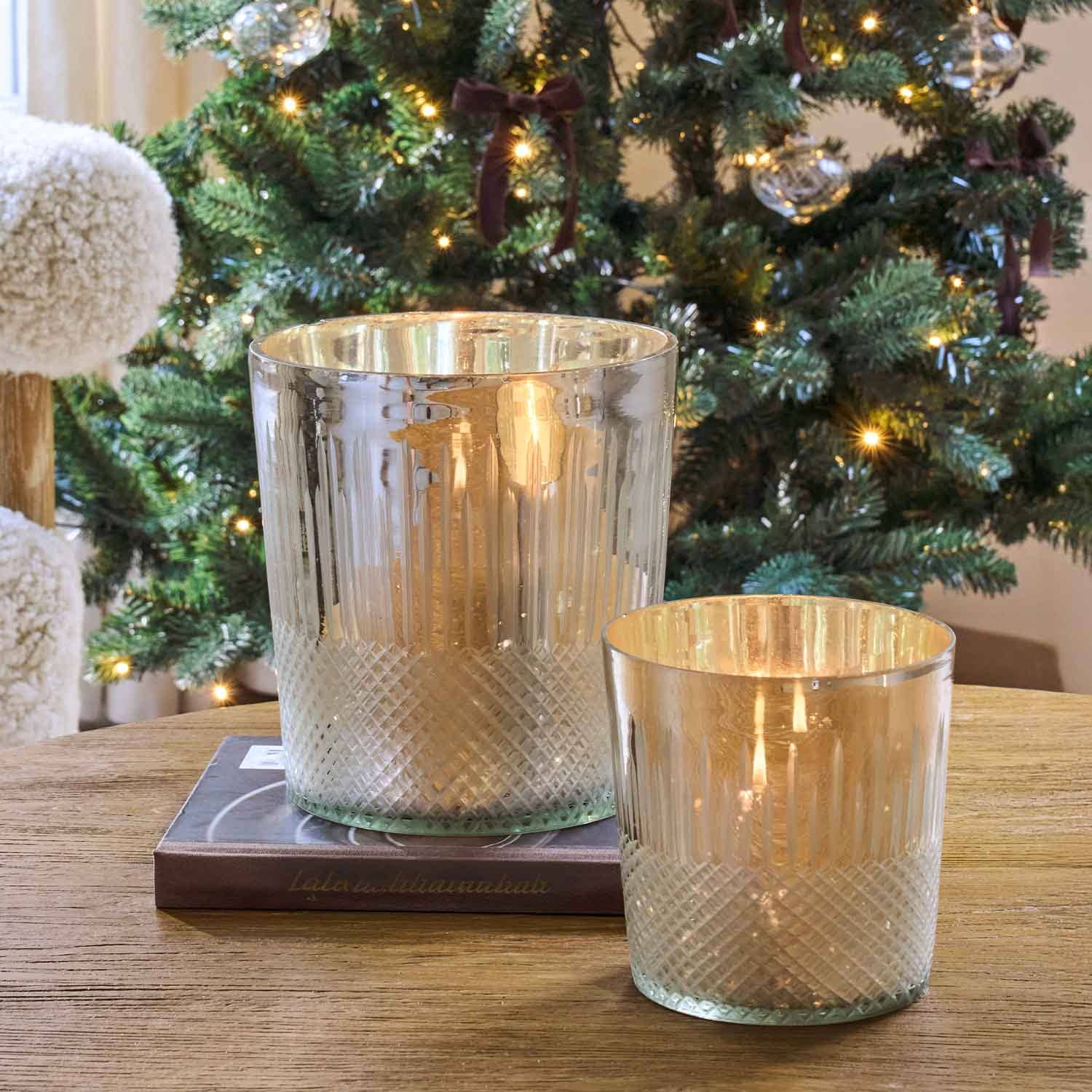 Twee glazen windlichten met metallic glans op houten tafel bij versierde kerstboom met lichtjes