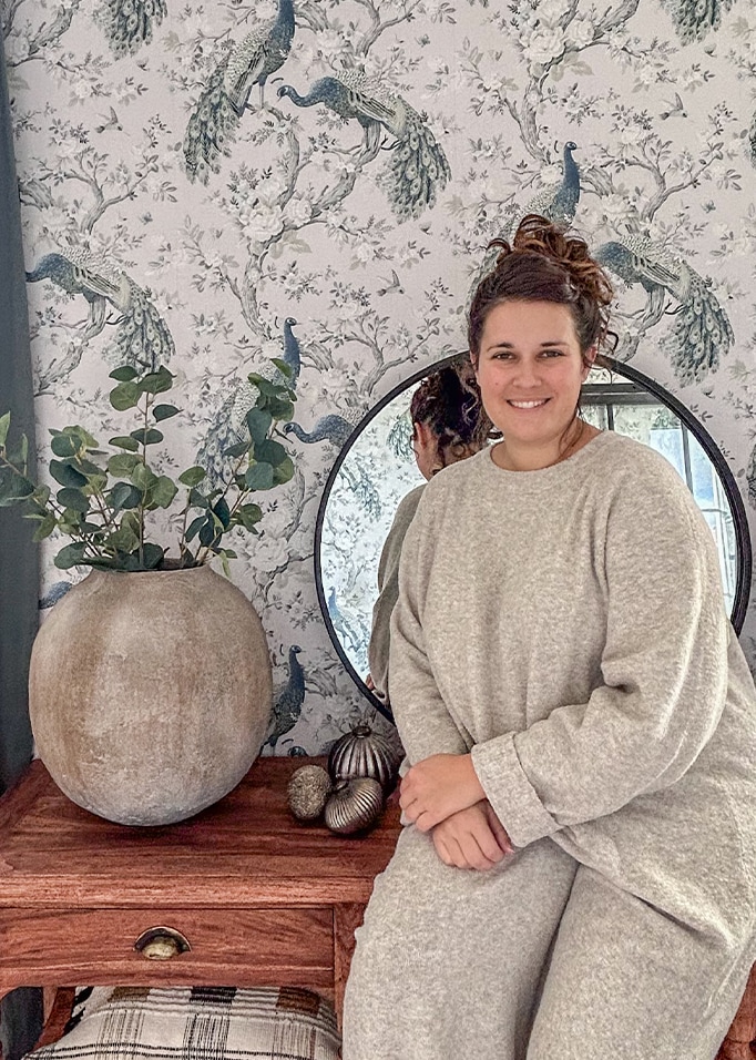 Vrouw in grijze trui zittend bij houten tafel met grote keramische vaas en spiegel voor een pauwenbehang